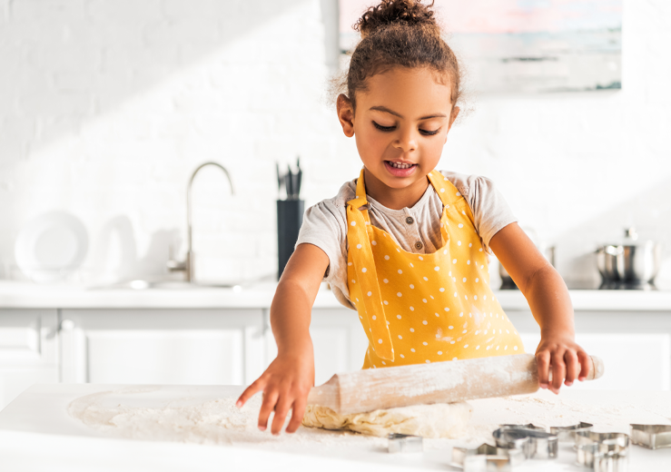 Female child in yellow apron using rolling pin on dough with cookie cutters on a white counter.