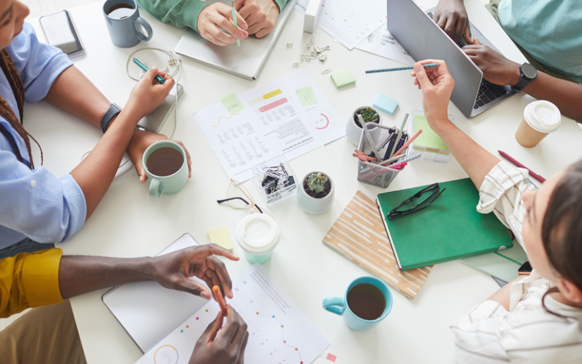 Close up of table during a meeting with coffee, papers, laptops, and diverse hands and arms showing.