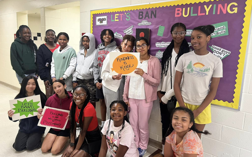 Large group of happy students holding colorful anti-bullying signs in front of an anti-bullying bulletin board inside of a school.