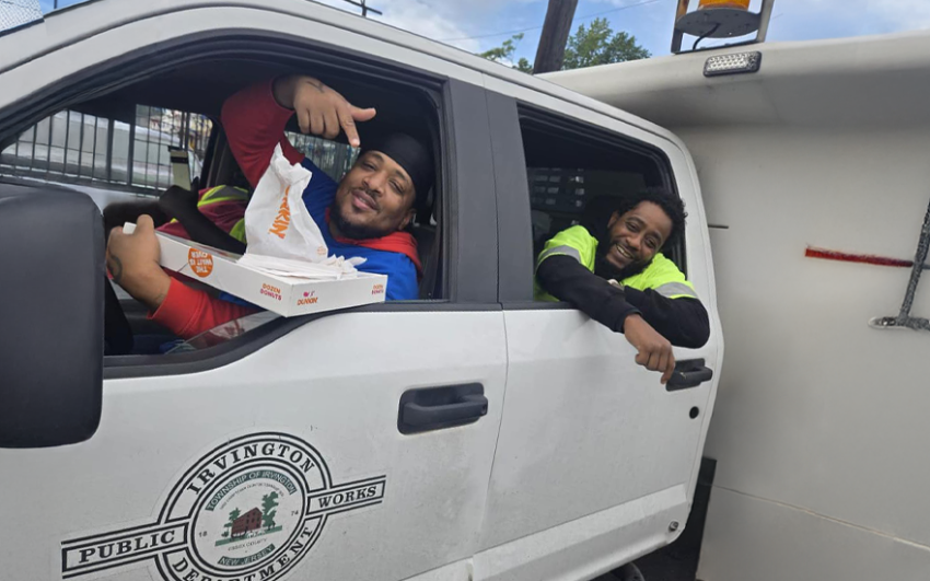 Two happy public works city workers in a white truck holding a box of donuts.