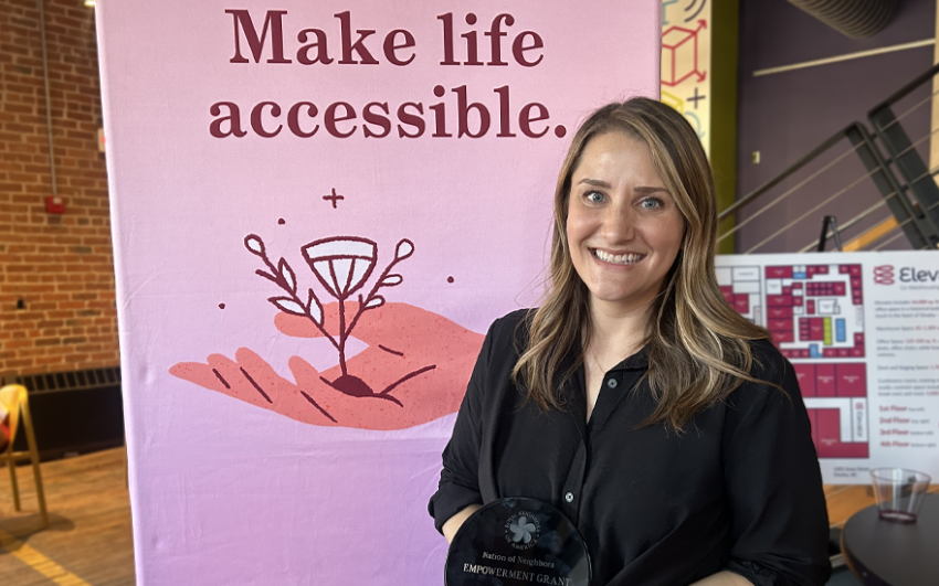 Smiling woman holding an award in front of a pink sign that says Mike life accessible.