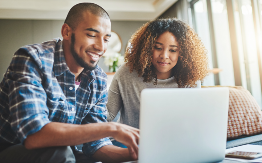 A happy couple sitting on a couch looking at a laptop.