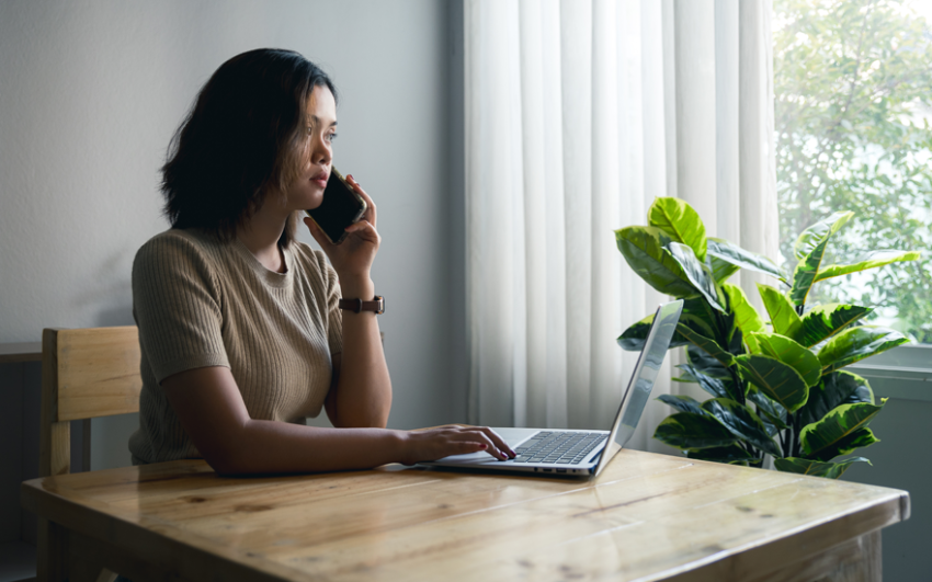 Concerned young woman on a phone at a table sitting in front of a laptop.