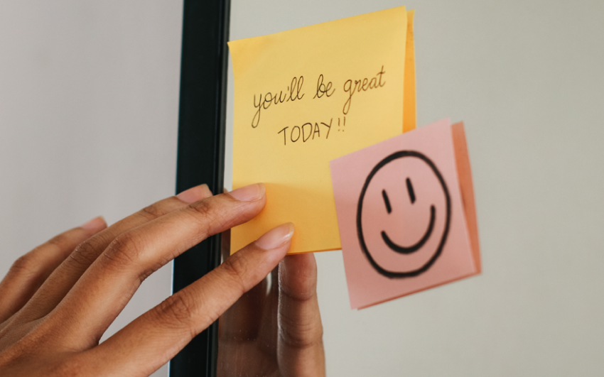 Close up of hand on yellow sticky note that says "You'll be great TODAY!" and a pink sticky note with a smiley face on it, both notes on a mirror.