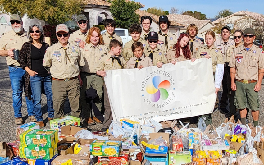 A large group of adults and kids from Girls Scouts and Scouting America with a Royal Neighbors banner outside with dozens of bags and boxes of food donations.